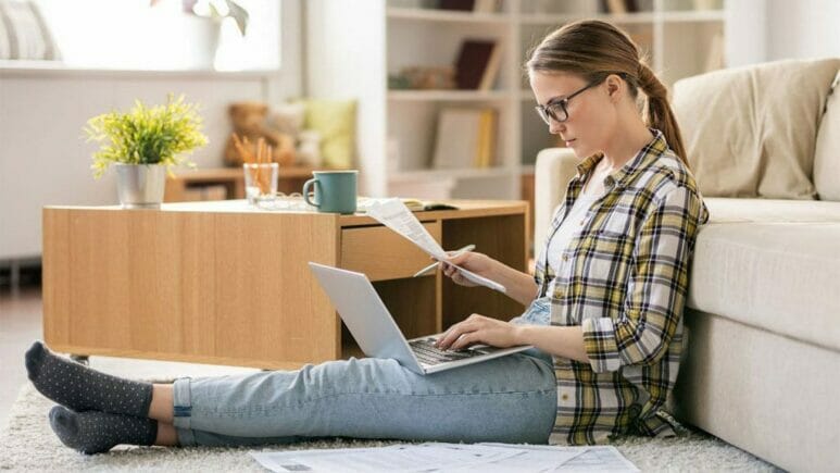 woman sitting on carpet in front of couch looking at computer and holding papers