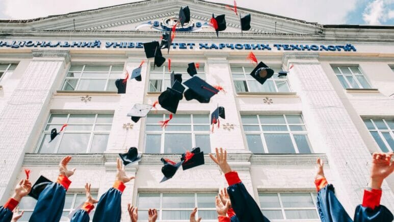 takingschooldistricts-16-9 school graduates standing in front of school throwing their graduation caps in the air